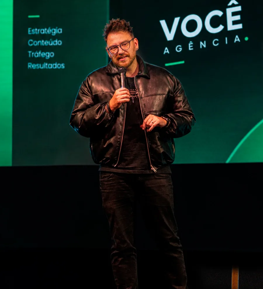 Abraão Salmoria falando num palco com a marca Você Agência no telão